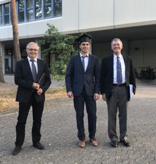 The photo shows Prof. Lenz, Peter Schwab and Prof. Meyer-Wegener after the doctoral examination. They are outside, in front of the entrance to the Computer Science Tower. All three men stand next to each other and wear casual dark suits. Peter Schwab is in the middle and wears an academic hat. All three smile at the camera.