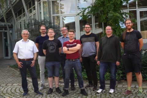 This is a group photo. Eight men in casual late-summer clothing stand next to each other outside in front of the training center. They are smiling at the camera.