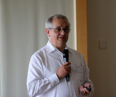 The picture shows Prof. Lenz in a close-up portrait. He wears a white shirt, rimless glasses and speaks into a hand-held microphone.