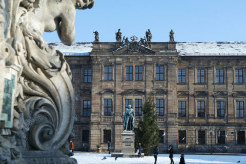 The picture shows a part of Erlangen's Schlossplatz. It's a winter shot; the sky is bright blue and there is a little snow. The photo shows the Erlangen palace which is now the seat of the university. In front of the castle we see the Margrave Monument, it represents the founder of the university, Margrave Friedrich von Brandenburg-Bayreuth. There is a very high christmas tree between the monument and the castle.