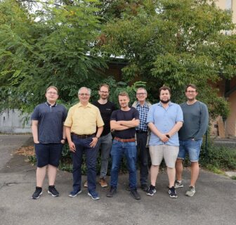 We can see a group photo. Seven men stand next to each other. There are two professors and five younger research assistants, around their late 20s or early 30s. The photo was taken outside in a city. The men stand in front of a large tree with a full canopy of leaves, behind which is a tall yellow building. It is late summer and some of the men are wearing short clothing. They look at the camera with a relaxed smile.