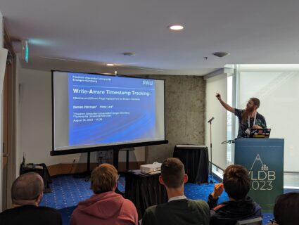 The picture was taken from the back of a conference room. The room is kept quite dark, there is a dark blue velor carpet on the floor. There are spectators sitting in the room, looking ahead. At the front of the room, on the right side, there is a lecturer standing behind a lectern. He points to a huge screen on the left side of the room. The title slide of a presentation is projected onto it.