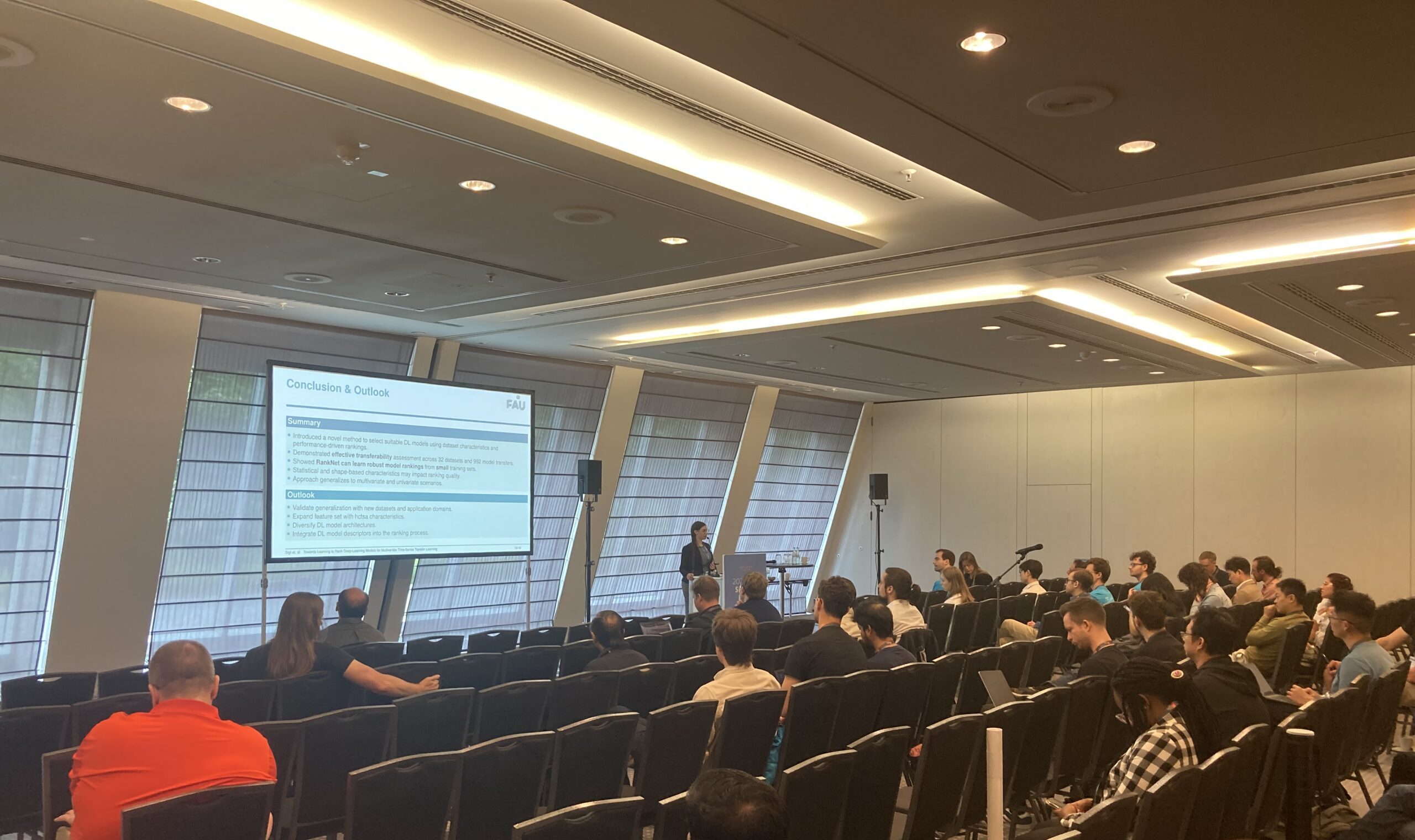 Melanie Sigl presents her research findings to an audience in the conference room. She is standing at the front of the room at the lectern, with the screen behind her. The rows in the conference room are sparsely filled.