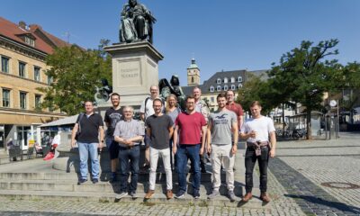 The picture shows 11 adults standing in Schweinfurt's central square. The group is standing directly in front of a bronze statue, with a church bell tower visible in the background. They are all wearing casual clothes and smiling at the camera.
